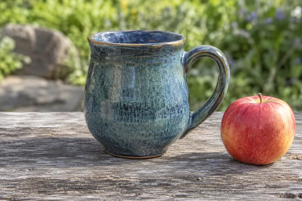 Une tasse à glaçure bleue, entièrement cadrée, se tient à côté d’une pomme rouge sur une surface en bois.